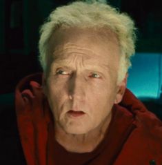 an older man with blonde hair and blue eyes stares at the camera while wearing a red hoodie