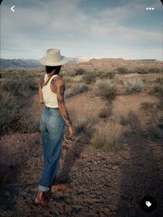 a woman in jeans and a hat is walking through the desert with her hand on her hip