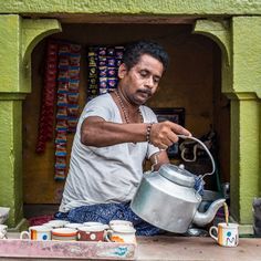 a man sitting on the ground pouring water from a pot into a teapot in front of a green building