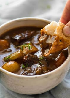 a hand dipping some food into a bowl filled with beef and potatoes in gravy