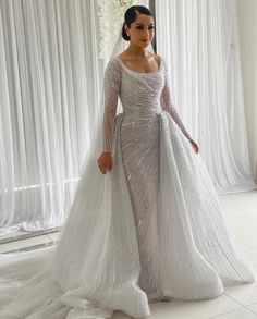 a woman standing in front of a window wearing a wedding dress with long sleeves and beading