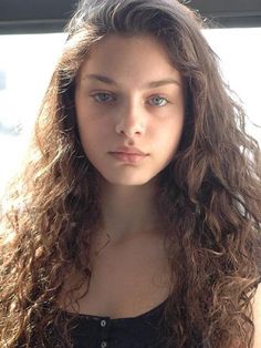 a young woman with long curly hair and blue eyes looking at the camera while standing in front of a window