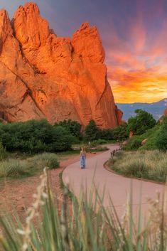 a woman is walking down a path in front of some mountains and trees at sunset