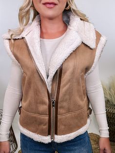 Camel Tan Suede Vest with Sherpa Trim Upgrade your winter wardrobe with our Camel Tan Suede Vest with Sherpa Trim. This chic vest is perfect for a stylish snow day outfit, offering a luxurious look and feel that will make you stand out. The soft suede material in a rich camel tan color is complemented by plush fur trim at the edges, giving it a high-end, sophisticated vibe. With a zipper front and side pockets, this vest combines practicality with elegance. The relaxed fit ensures comfort, makin Chic Brown Winter Vest, Fall Sleeveless Outerwear With Faux Fur Trim, Sleeveless Outerwear With Faux Fur Trim For Fall, Sleeveless Outerwear With Faux Fur Lining For Fall, Sleeveless Fall Outerwear With Faux Fur Lining, Cozy Winter Vest For Cold Weather, Trendy Winter Vest For Cold Weather, Brown Winter Vest Outerwear, Winter Beige Sleeveless Outerwear