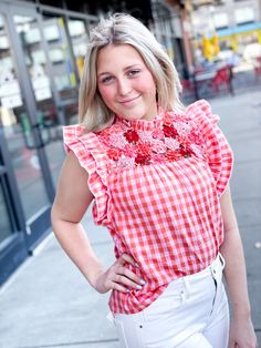 Blonde woman standing in a shopping center wearing a pink and orange gingham top with intricate flower details at the chest. The flowers are made from coordinating fabrics and woven together to create the chest/shoulder of the top. The shoulders feature of double ruffle. She has the top paired with white denim jeans. Orange Shades, White Denim Jeans, Coordinating Fabrics, Gingham Print