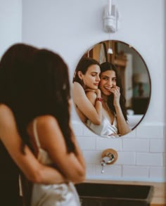 two women standing in front of a mirror looking at each other's faces and smiling