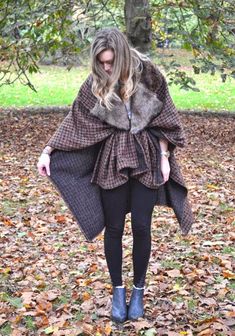 a woman standing in the leaves wearing a cape and boots with her hands on her hips