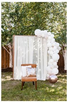 a chair and balloon arch in the grass at a backyard party with white balloons on it