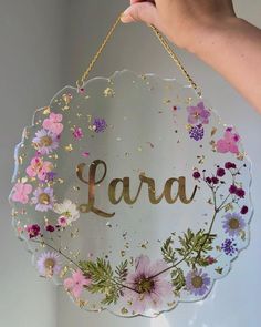 a hand holding a glass sign with flowers on it that says lara in gold lettering