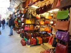 many purses and handbags are on display in a market area with people walking by