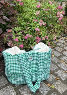 a green and white checkered bag sitting on the ground next to some bushes with pink flowers