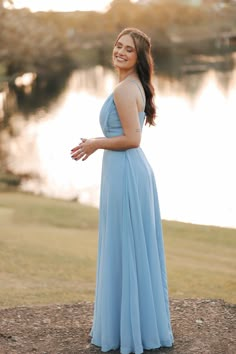 a woman in a long blue dress standing on the edge of a lake and smiling