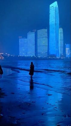 two people walking on the beach in front of tall buildings at night with blue lights