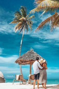 a man and woman standing on the beach under a palm tree next to the ocean