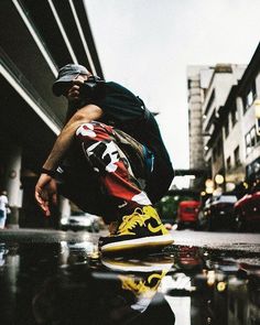 a man sitting on top of a skateboard next to a wet street with buildings in the background