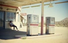 a person sitting on a bench in front of a gas station with two old fashioned machines