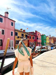 a woman walking down a sidewalk next to colorful buildings and boats in the water on a sunny day