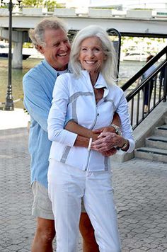 an older man and woman standing next to each other in front of a bridge with their arms around each other