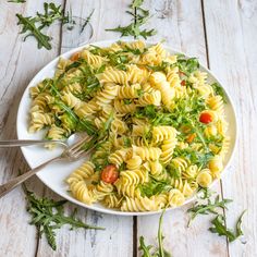 a white bowl filled with pasta and greens on top of a wooden table next to a fork