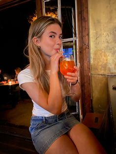 a woman sitting on a window sill drinking from a tall glass in front of her