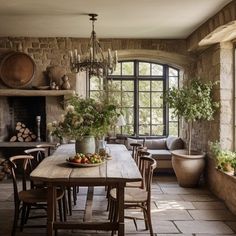 a rustic dining room with stone walls and flooring is furnished with wood furniture, potted plants, and an old - fashioned chandelier