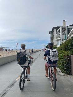 two people riding bikes down a sidewalk near the beach