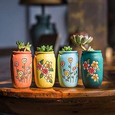 four painted flower pots sitting on top of a wooden table next to a vase with succulents in it