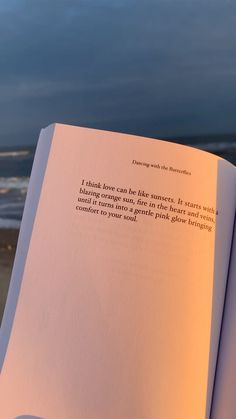 an open book sitting on top of a beach next to the ocean with a sunset in the background