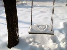 a swing with a heart drawn on it in the snow next to a tree trunk