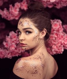 a woman with glitter on her face and body in front of pink flowers, looking at the camera