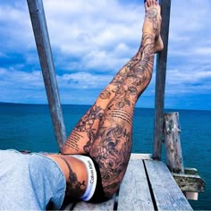 a man laying on top of a wooden dock next to the ocean with tattoos on his legs