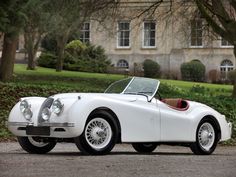 an old fashioned white sports car parked in front of a large building with trees and bushes