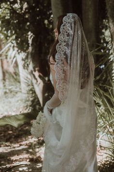 a woman in a wedding dress and veil looking at the ground with trees behind her