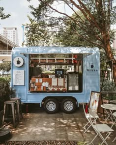 a blue food truck parked on the side of a road next to trees and chairs