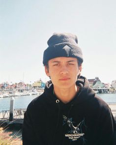 a young man in a black hoodie is standing by the water and looking at the camera