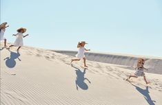 four women in white dresses and straw hats run across the sand dunes on a sunny day