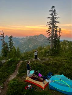 two people standing on top of a mountain next to tents and backpacks at sunset