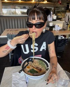 a woman eating noodles with chopsticks at a table in a restaurant, while wearing sunglasses