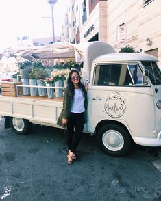 a woman standing in front of an old truck with flowers on the bed and potted plants behind it