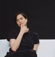 a woman sitting on top of a white couch next to a black wall and holding her hand under her chin