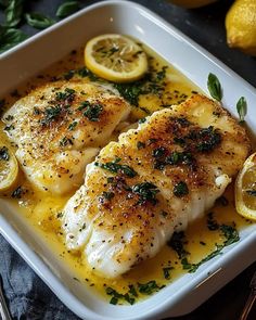 two fish fillets in a white dish with lemons and herbs on the side