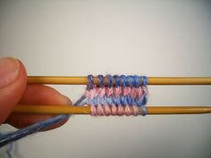 a hand holding two knitting needles with blue and pink yarn on them next to an orange needle