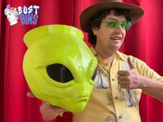 a man in a hat and glasses holding a yellow alien mask