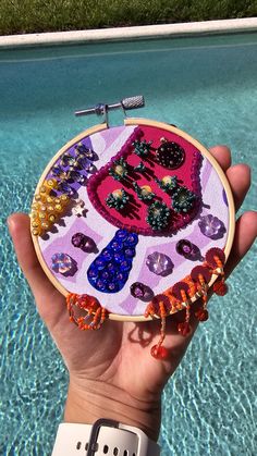 someone is holding up a small embroidery project in their hand near a pool with blue water