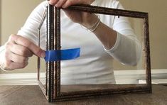 a woman holding up a mirror with a blue object in it's frame on top of a wooden table