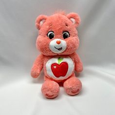 a pink teddy bear with an apple on it's chest sitting against a white background