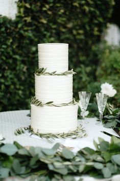 a white wedding cake sitting on top of a table next to greenery and wine glasses