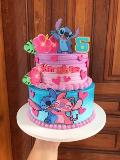 a hand holding a three tiered cake decorated with cartoon characters and flowers on it