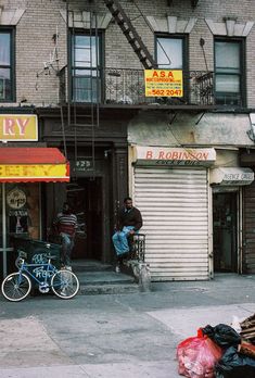 two people sitting on the sidewalk in front of a building with an awning and fire escape