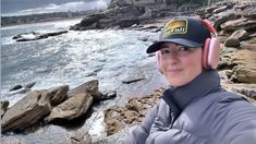 a woman wearing headphones standing next to the ocean with rocks and houses in the background